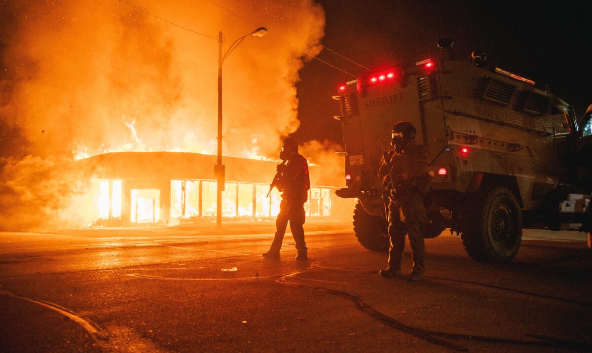 Một chiếc xe bọc thép của cảnh sát đang tuần tra ở một ngã tư trong lúc một tòa nhà bị đốt cháy bởi những người bạo loạn ở thành phố Kenosha, bang Wisconsin, vào ngày 24/8/2020. (Ảnh Brandon Bell / Getty Images)