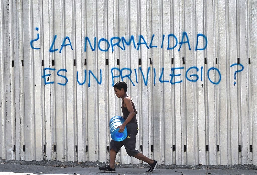 Một cậu bé khiêng theo một bình đựng nước khi đi ngang qua một bức vẽ graffiti viết "Sự bình thường có phải là một đặc quyền?" trong một thời kỳ thiếu điện mới ở Venezuela, tại Đại lộ Fuerzas Armadas ở Caracas vào ngày 31/3/2019. (Ảnh Federico Parra/ AFP qua Getty Images)