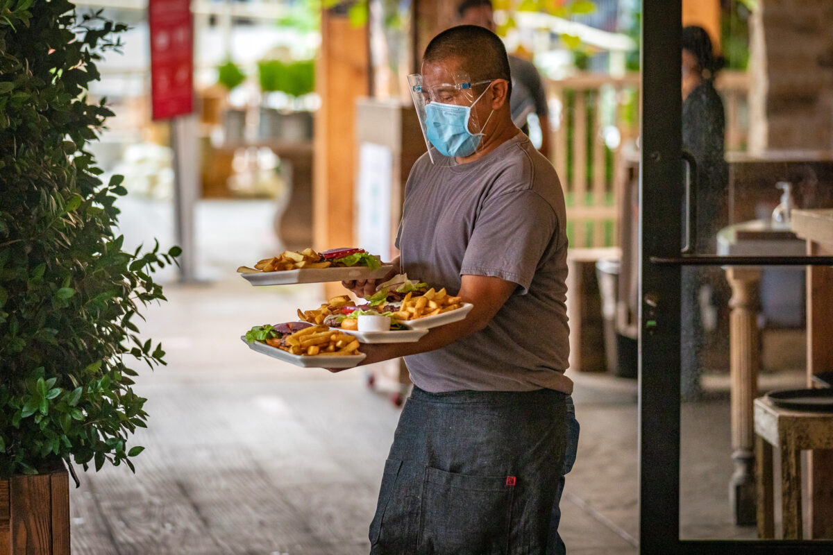 Một người phục vụ đeo đồ bảo hộ trên khuôn mặt, giữ thăng bằng các đĩa thức ăn trên tay khi  giao cho thực  khách tại một nhà hàng ở Newport Beach, California, ngày 9 tháng 9 năm 2020. (Ảnh John Fredricks / The Epoch Times)