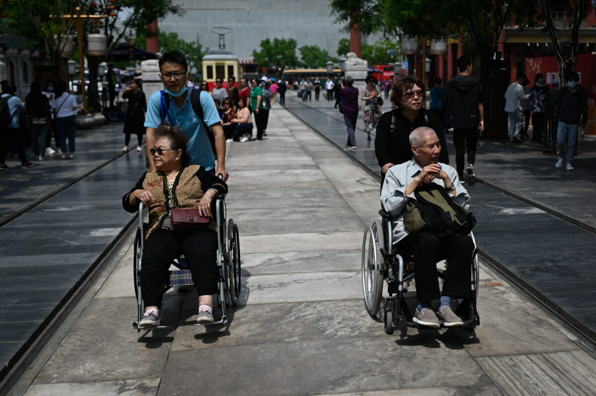 Một bác trai và một bác gái lớn tuổi được đẩy trên xe lăn dọc theo một con phố ở Bắc Kinh hôm 11/05/2021. (Ảnh: Wang Zhao/AFP qua Getty Images)