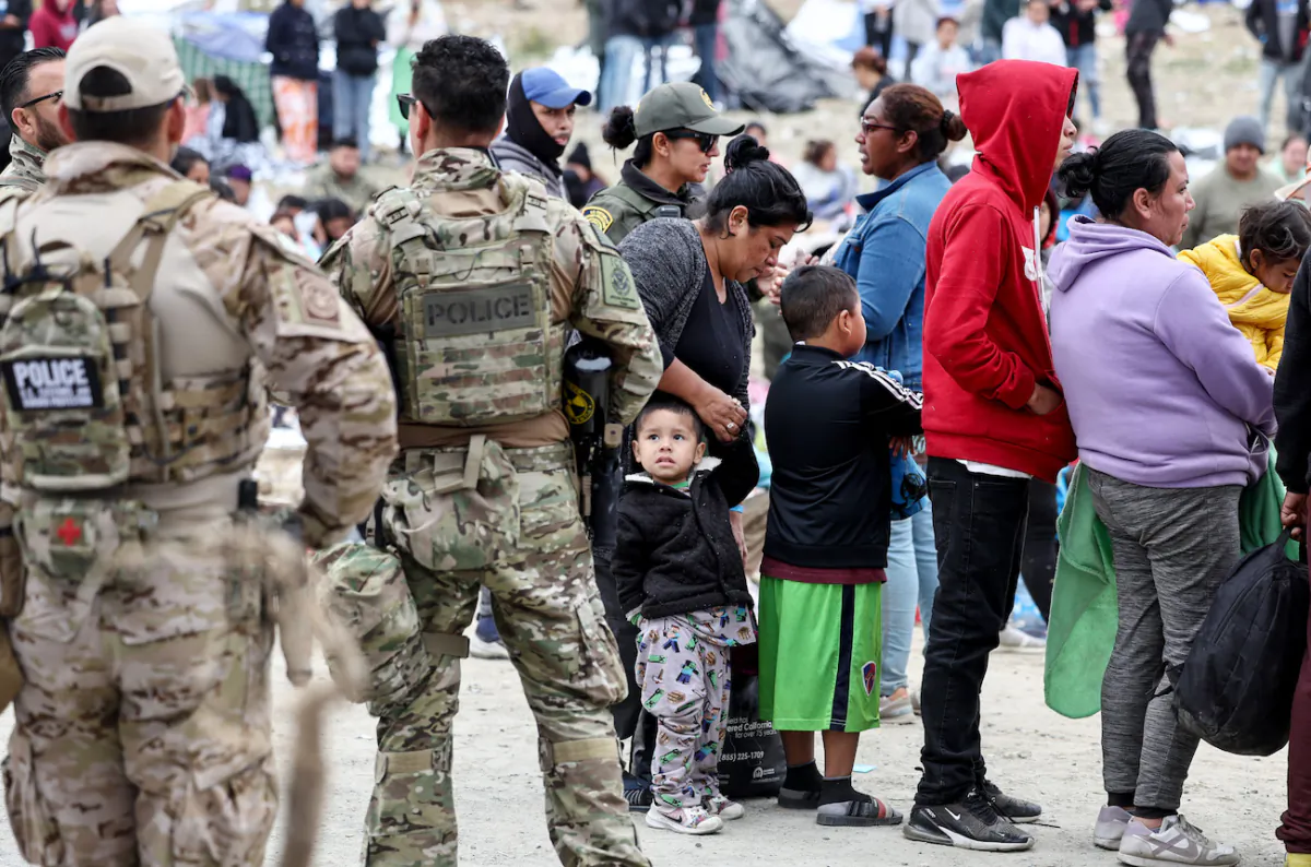 Các nhân viên và quan chức chấp pháp liên bang đang theo dõi trong khi những người nhập cư xếp hàng để được chở đi từ một trại tạm bợ giữa các bức tường biên giới Hoa Kỳ-Mexico ở San Diego hôm 13/05/2023. (Ảnh: Mario Tama/Getty Images)