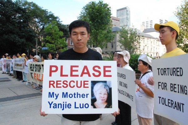 Anh Phạm Văn Thác (Wenta Fan) cầm một tấm biểu ngữ có bức ảnh chân dung của mẹ mình, bà Lạc Diễm Kiệt (Yanjie Luo), người đã bị kết án 13 năm tù ở Trung Quốc vì tu luyện Pháp Luân Công vào năm 2011, bên ngoài Tòa thị chính Toronto vào ngày 25/08/2016. (Ảnh: Yi Ling/The Epoch Times)