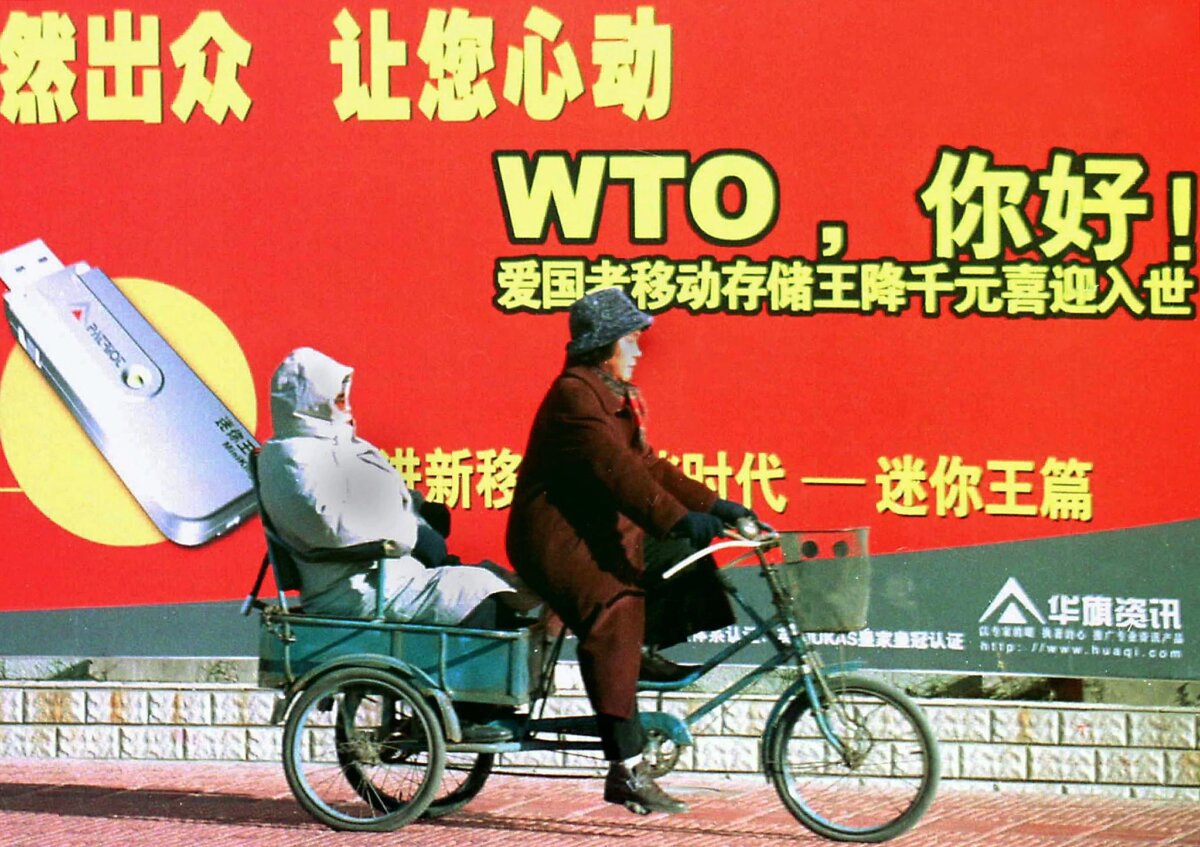 Một cặp vợ chồng người Trung Quốc đi xe ba bánh ngang qua bảng quảng cáo chào mừng quốc gia này trở thành thành viên Tổ chức Thương mại Thế giới (WHO), dọc một con phố ở Bắc Kinh ngày 23/12/2001. (Ảnh: AFP qua Getty Images)